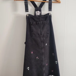 Svart jeansklänning med broderade blommor - Trendig svart jeansklänning med justerbara axelband och broderade blommor i olika färger. Klänningen har en stor ficka framtill och en avslappnad passform. 