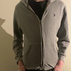 Grå zip hoodie från Ralph lauren - Säljer en stilren grå huvtröja från Polo Ralph Lauren. Modellen är 170 men den är lite för liten så den passar någon lite kortare. Hoodien är i relativt bra skick därav priset