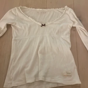 Vit ribbad topp från Odd Molly - Säljer en söt vit ribbad topp från Odd Molly med en liten vinröd rosett vid halsringningen. Toppen har långa ärmar och en fin  detalj på ärmen. Storlek 1