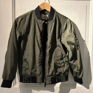 Grön bomberjacka från New York Studios - Snygg grön bomberjacka från New York Studios med svarta ribbade muddar och dragkedja. Jackan har en cool detalj med text på fållen och en ficka med dragkedja på ärmen. Perfekt för en stilren look.