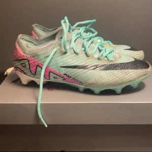 Nike fotbollsskor  - Snygga Nike fotbollsskor i mintgrön med rosa och svarta detaljer. Skorna har snörning och en modern design med tydliga logotyper på sidorna. Perfekta för fotbollsplanen med en stilren look.