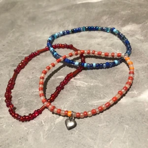 Färgglada pärlarmband - Tre färgglada pärlarmband i olika nyanser. Ett armband är blått med små pärlor, ett är rött och det tredje är orange med ett litet silverhjärta som detalj. Perfekta för att lägga till en färgklick till din outfit! Berlocken kostar 10 kr armbanden 40kr så alla tsm blir 90kr