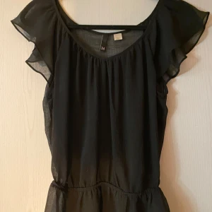 Svart topp med volangärmar - Snygg svart tröja med volangärmar och rund halsringning. Perfekt för en stilren look. Toppen har en luftig passform och är tillverkad i ett lätt material.