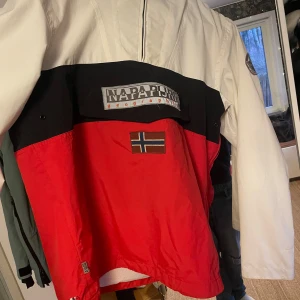 Röd och vit jacka från Napapijri - Säljer en snygg röd och vit jacka från Napapijri med svart detalj och norsk flagga på framsidan. Jackan har en dragkedja och en stor ficka framtill. Perfekt för kyliga dagar med sin stilrena design och även i skidbacken. Köp så postar jag samma dag:)