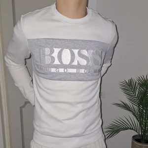 Vit tröja från Hugo Boss - Säljer en stilren vit tröja från Hugo Boss med ett grått parti över bröstet där märkesnamnet är tryckt i stort. Tröjan har långa ärmar och en rund halsringning. Aldrig använd. Perfekt för en avslappnad och trendig look.