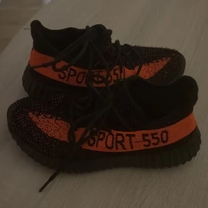 Svarta och orange sneakers - Snygga svarta sneakers med orange detaljer och texten 'SPORT 550' på sidan. Skorna har en stickad ovandel och snörning för en bekväm passform. Perfekta för en sportig look.