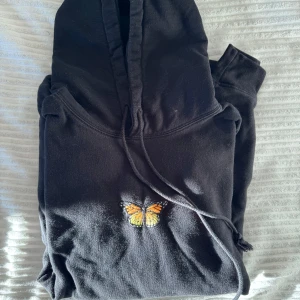 Svart hoodie med fjärilsbroderi från Brandy Melville - Säljer en svart hoodie från Brandy Melville med ett snyggt broderi av en fjäril på bröstet. Hoodien har en klassisk design med dragsko i huvan och en stor ficka framtill. Perfekt för en avslappnad stil.