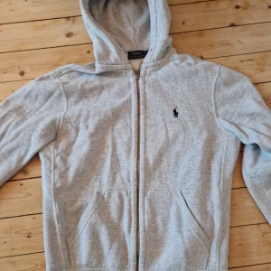 Grå hoodie från Ralph Lauren - Säljer en stilren grå hoodie från Ralph Lauren med dragkedja och klassisk logga på bröstet. Perfekt för en avslappnad look. Den har en bekväm passform och praktiska fickor framtill.