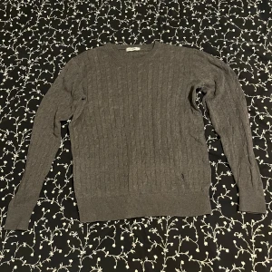 Grå Kabelstickad tröja - Säljer en grå kabelstickad tröja från J.Lindeberg. Perfekt för kyligare dagar. Den har en diskret logga framtill.