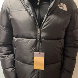 Svart dunjacka från The North Face - Säljer en svart dunjacka från The North Face med dragkedja och hög krage. Jackan har en pufferdesign och är perfekt för kalla dagar. Den har en logotyp på bröstet och praktiska fickor med dragkedja. PRISET KAN DISKUTERAS!!