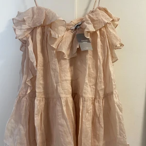 Beige volangklänning från H&M Edition - Superfin kortklänning i ljus beige från H&M Edition. Klänningen har tunna axelband, volanger upptill och ett luftigt, flowy tyg i linne-look. Perfekt för varma dagar när du vill känna dig både söt och bekväm.