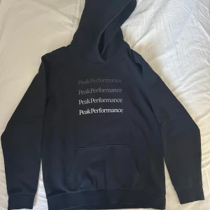 Svart hoodie från Peak Performance - Svart hoodie från Peak Performance med tryckt logga i flera nyanser på bröstet. Klassisk känguruficka framtill och stor huva. Perfekt för dig som gillar en stilren och sportig look. Mjuk och skön bomullsblandning. Hoodien är i mycket bra skick, nästan i nyskick så den använts sparsamt. Pris går att diskutera vid smidig och snabb affär. Har du frågor är det bara att höra av dig😊