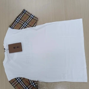 Vit Burberry t-shirt med rutiga ärmar - Vit t-shirt från Burberry med klassiska rutiga korta ärmar i beige, svart, rött och vitt. Stor svart logga framtill med texten 'BURBERRY LONDON ENGLAND'. Skön passform och stilren design, perfekt för dig som gillar exklusiva streetwear-vibbar.
