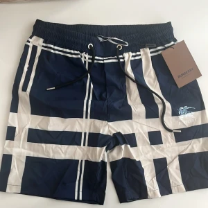 Blåvita Burberry badshorts med rutor - Snygga badshorts eller sommarshorts från Burberry i marinblått och vitt med klassiskt rutmönster. Shortsen har elastisk midja med svart snörning och Burberry-logga framtill. Perfekta för stranden eller poolen. Storleken passar. XL ELLER L
