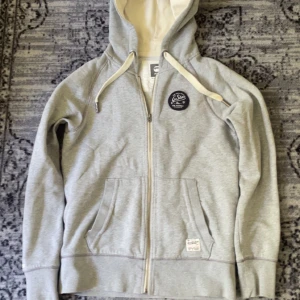 Grå hoodie från G-Star Raw med dragkedja - Snygg ljusgrå hoodie från G-Star Raw med hel dragkedja, huva med snörning och två stora fickor framtill. Insidan av huvan är beige/vit och tröjan har en svart patch på bröstet. 