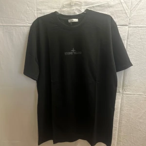 Svart Stone Island t-shirt med tryck - Svart t-shirt från Stone Island med klassisk logga tryckt på bröstet och stort grafiskt tryck på ryggen. Skön passform och rund halsringning, perfekt till jeans eller shorts. Materialet känns mjukt och är troligtvis bomull.