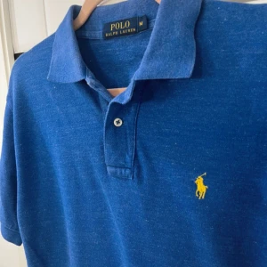 Blå pikétröja från Polo Ralph Lauren - Klassisk blå pikétröja från Polo Ralph Lauren med korta ärmar och gul broderad logga på bröstet. Tröjan har krage och knäppning framtill. Perfekt för en stilren och avslappnad look.