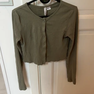 Olivgrön croppad kofta från H&M Divided - Snygg olivgrön croppad kofta från H&M Divided i mjuk bomull. Koftan har långa ärmar, rund halsringning och knappar hela vägen framtill. Perfekt att slänga över en topp för en chill look.