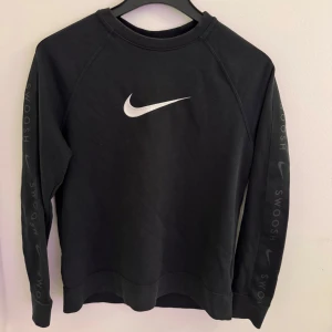 Svart Nike sweatshirt med Swoosh-logga - Svart sweatshirt från Nike med stor vit Swoosh-logga på bröstet och texten 'Swoosh' längs båda ärmarna. Klassisk rund halsringning och långa ärmar. Perfekt för en sportig och avslappnad stil. Den är i storlek Xl men tycker att den verkar vara mindre jag har S och brukar ha den som en oversized tröja