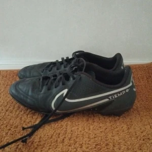 Nike Tiempo svarta fotbollsskor - Svarta Nike Tiempo fotbollsskor med vita detaljer och klassisk swoosh-logga på sidan. Skorna har snörning och dobbar för bra grepp på planen. Tillverkade i syntetmaterial med rund tå och låg profil. Perfekta för dig som vill ha stilrena och funktionella skor till matchen.