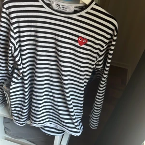 Randig långärmad tröja Comme des Garçons - Säljer en svartvit randig långärmad tröja från Comme des Garçons Play med det ikoniska röda hjärtat broderat på bröstet. Tröjan är i mjuk bomull och har en klassisk passform med rund hals. Perfekt för dig som gillar stilrena statement-plagg.