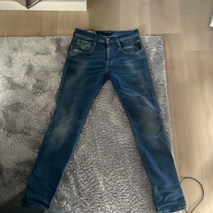 Replay Anbass blå jeans, W28 L30 - Säljer ett par snygga Replay Anbass jeans i blå tvätt med coola slitningar och klassiska fem fickor. Jeansen har smal passform och tydliga kontrastsömmar. Märkespatch bak i midjan och dragkedjegylf. Perfekta för dig som gillar stilrena jeans med edge.
