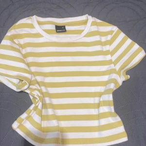 Randig topp från Gina Tricot - Superfin topp från Gina Tricot med breda gula och vita ränder. Modellen är kortärmad med rund halsringning och har en klassisk, avslappnad passform. Perfekt till jeans eller kjol för en fräsch och enkel look.