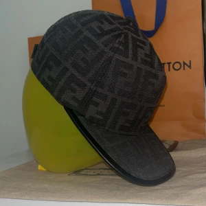Svart Fendi keps med FF-mönster - Snygg svart keps från Fendi med det klassiska FF-logomönstret över hela kepsen. Kepsen har böjd skärm med svart kant och justerbar rem baktill. Tillverkad i ett slitstarkt tyg som känns lyxigt och trendigt. Perfekt för dig som vill sticka ut med designerstil.