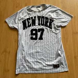 Vit randig t-shirt New York FB Sister - Vit t-shirt från FB Sister med svarta tunna ränder och stor svart text 'NEW YORK 97' på bröstet. Sportig collegekänsla. Skön att ha på sig nu under sommaren, kan finnas lite defekter men inget man har stört sig på 🌸
