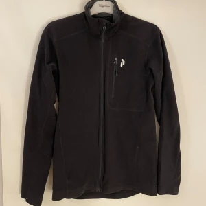 Svart fleecejacka Peak Performance S - Svart fleecejacka från Peak Performance med hel dragkedja och hög krage. Jackan har en bröstficka med dragkedja och diskret logga på bröstet. Perfekt för lager-på-lager och chill dagar. Långärmad och i mjukt fleecematerial.