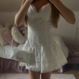 Vit klänning med spetsdetaljer - Supersöt vit klänning med broderade blommor och spetsdetaljer vid v-ringningen. Klänningen har en figurnära överdel och volangig nederdel. Perfekt för sommaren, ALDRIG ANVÄND!