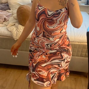 Färgstark swirl kortklänning - Säljer en kort klänning med swirlmönster i orange, brun, vit och svart. Klänningen har smala axelband, figurnära passform och snygg rynkdetalj på sidan. Perfekt för dig som gillar att sticka ut med färg och mönster!