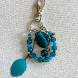 Blå nyckelring/ bag charm med fjäder - Unik nyckelring med blå glaspärlor, stora dekorativa pärlor och en cool blå fjäderdetalj. Silverfärgad karbinhake gör det enkelt att fästa på väskan eller använda som smycke. Perfekt accessoar för dig som vill sticka ut med färg och personlighet. 
