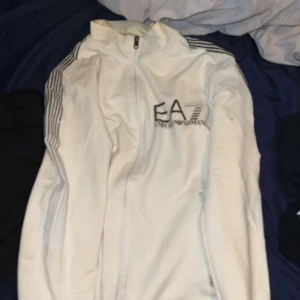 Vit EA7 vindjacka med dragkedja - Snygg vit vindjacka från EA7 Emporio Armani med hel dragkedja framtill och svarta ränder på axlarna. Jackan har hög krage och EA7-logga på bröstet. Perfekt för dig som gillar sportig och clean stil.