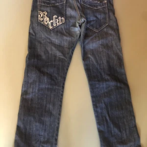 Mörkblå jeans med broderad bakficka - Snygga mörkblå jeans med rak passform och coolt broderi med texten 'Rockin' på ena bakfickan. Klassisk femficksmodell i jeansmaterial med kontrastsömmar. Perfekta för dig som vill ha en unik detalj på dina jeans.