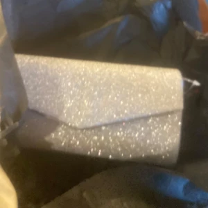 Glittrig silver kuvertväska - Säljer en snygg kuvertväska i silver med glittrig yta. Väskan har en stilren, avlång form och stängs med ett lock. Perfekt accessoar för att lyfta din outfit med extra bling.
