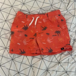 Röda badshorts  - Snygga röda badshorts från Freefly med tropiskt palmmönster i blått, vitt och gult. Shortsen har elastisk midja med vit snörning, sidfickor och en bakficka med knapp. Perfekta för stranden eller poolen, med meshfoder för extra komfort. 
