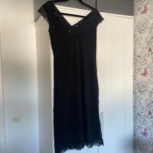 Llångklänning med paljetter - Svart långklänning från KappAhl med v-ringning och dekorativa paljetter längs halsringningen. Klänningen har korta, transparenta spetsärmar och spetsdetalj vid fållen. 