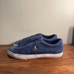 Blå mockasneakers från Polo Ralph Lauren - Snygga blå sneakers i mocka från Polo Ralph Lauren med klassisk siluett och vit platt sula. Skorna har snörning och den ikoniska broderade Polo-loggan i guld på sidan. Perfekta för en clean och stilren look.