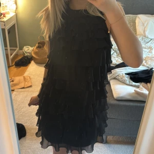 Svart volangklänning med öppen rygg - Supersnygg svart kortklänning med flera lager volanger och öppen rygg. Klänningen är ärmlös och har en luftig känsla, perfekt för dig som vill sticka ut. Materialet känns lätt och flowy, och ryggen har en cool cut-out detalj.