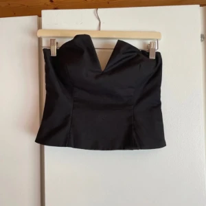 Svart tubtopp med v-ringad kant från Kappahl - Säljer en svart tubtopp med snygg v-ringad kant upptill. Toppen är axelbandslös och har smockade partier baktill för extra komfort och passform. Perfekt att styla med höga jeans eller kjol för en trendig look. Nypris:500