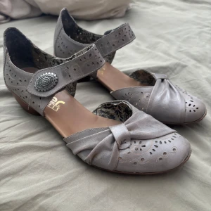 Grå ballerinaskor med klack från Rieker - Snygga grå ballerinaskor från Rieker med låg klack, dekorativt spänne och utskurna mönster. Skorna har rund tå, rosett framtill och är gjorda i skinn. Perfekta för dig som gillar en feminin och vintageinspirerad stil.