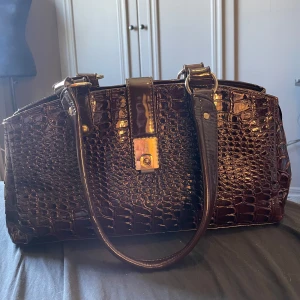 Mörkbrun croco handväska med dragkedja - Snygg mörkbrun/ vinröd handväska i croco-mönstrat skinnimitation med guldiga metalldetaljer. Väskan har två handtag, dragkedja på baksidan och ett stilrent spänne framtill. Perfekt för dig som vill ha en classy och trendig look.