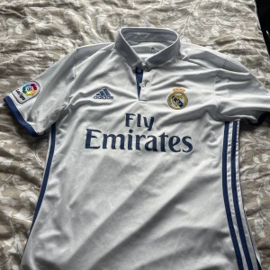 Real Madrid Adidas fotbollströja M - Snygg Real Madrid fotbollströja från Adidas med klassisk krage och korta ärmar. Vit med blå detaljer, klubbmärke och Fly Emirates-tryck på bröstet. La Liga-märke på ärmen och tillverkad i lätt och ventilerande ClimaCool-material.