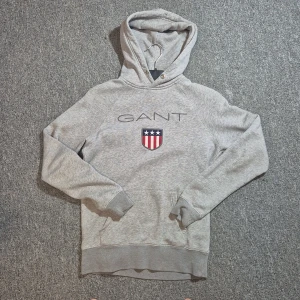 Grå hoodie från GANT  - Snygg grå hoodie från GANT i storlek XS. Tröjan har broderad GANT-logga och ett sköldmärke med stjärnor och ränder på bröstet. Klassisk huva och känguruficka framtill. Mjuk och skön bomullsblandning, perfekt för chill dagar.