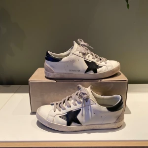 Golden Goose vita sneakers med svart stjärna - Golden Goose sneakers i vitt skinn med svart stjärna på sidan och svart häl. Grå snörning och platt sula. Klassisk låg modell med rund tå och coola detaljer. Perfekta för dig som gillar streetstyle och vill ha en unik touch på outfiten.    STR 40 - Nypris 4500+