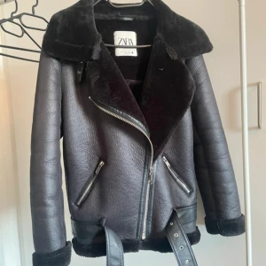 Svart jacka från Zara - Säljer en svart Zara jacka i storlek XS. Jackan har bred krage, dragkedja i sned modell, två fickor med dragkedja och bälte nertill. Insidan och kragen är fodrad med mjuk teddy. Perfekt för kalla dagar och riktigt snygg till jeans.