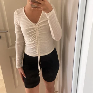 Vit ribbad topp med snörning från Sister - Vit långärmad topp från Sister i ribbat material. Djup v-ringning och snygg snörning framtill som ger en rynkad effekt. Croppad passform som sitter tajt och framhäver midjan. Perfekt till jeans eller kjol för en trendig look.