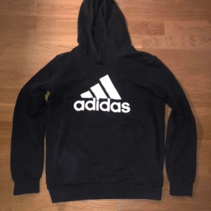 Svart Adidas hoodie med stor logga - Svart hoodie från Adidas med stor vit logga på bröstet. Klassisk känguruficka framtill, huva och långa ärmar. Perfekt för en sportig och avslappnad stil. Aldrig använd!!!