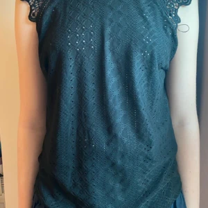 Mörkgrön spetsblus med knyt i nacken - Supersöt svart blus med spetsdetaljer vid axlarna och ett fint knyt i nacken. Blusen har ett diskret hålmönster och är ärmlös, perfekt för en trendig och luftig look. Passar dig som vill ha något extra till outfiten.